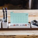 Calendario mensile appoggiato su una scrivania accanto a oggetti di uso quotidiano.